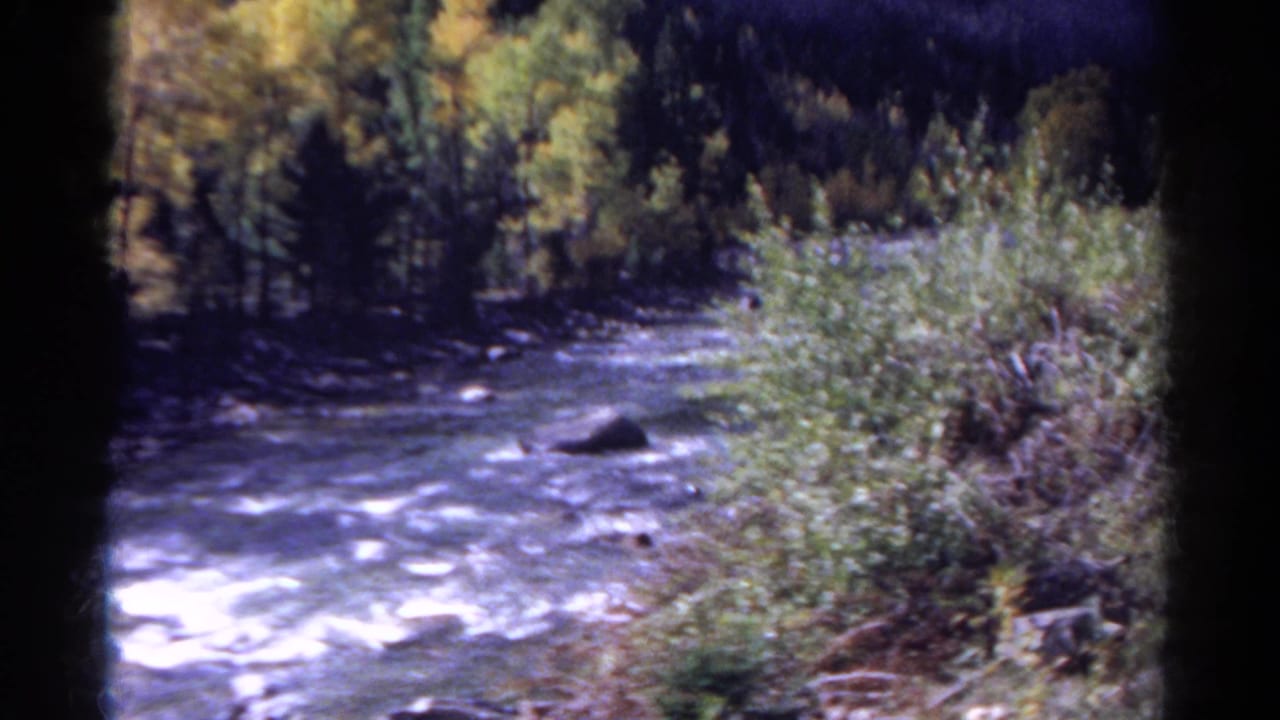Rushing river bordered by trees and brush, seen through dark 8mm film-frame edges.