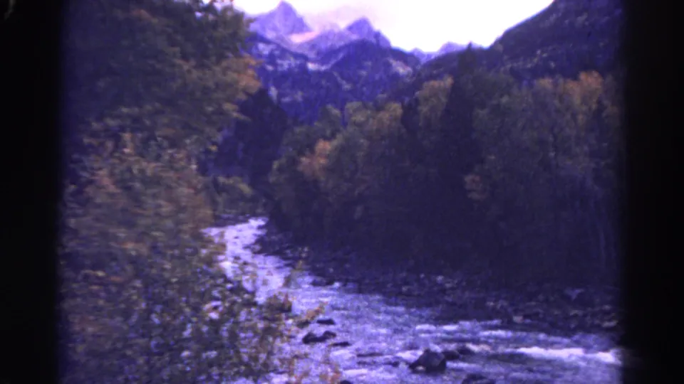 1967 Colorado home-movie frame showing a forested creek, pale rushing water, rocks, and mountain peaks in the distance.