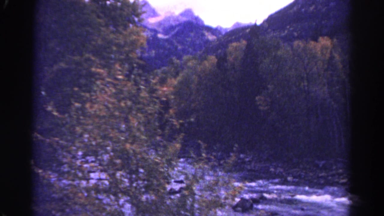 1967 Colorado 8mm frame showing a rocky creek between tree-lined banks with dark mountain slopes and bright sky beyond.