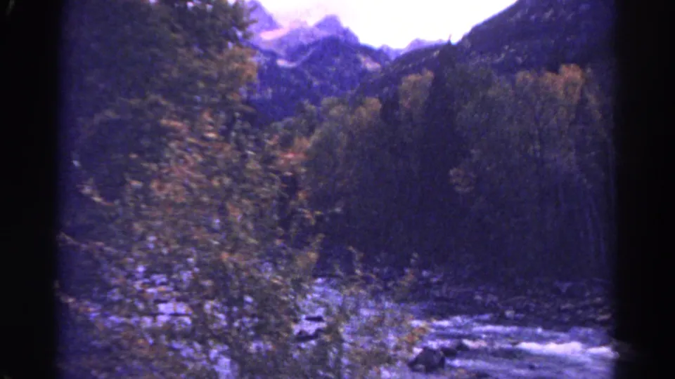 1967 Colorado 8mm frame showing a rocky creek between tree-lined banks with dark mountain slopes and bright sky beyond.