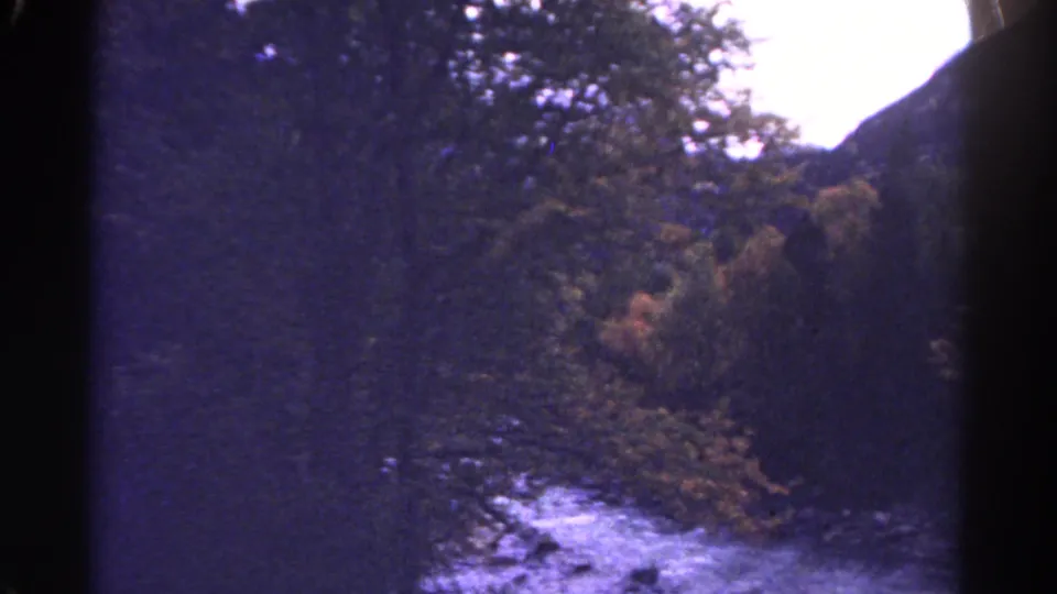 1967 Colorado 8mm frame with flowing water, dense roadside trees, rocks, and a dark mountain slope edging the scene.