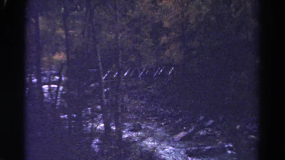 1967 Colorado 8mm frame showing a shadowed creek edge, rocks, trees, and what appears to be a small fence or rail near the water.