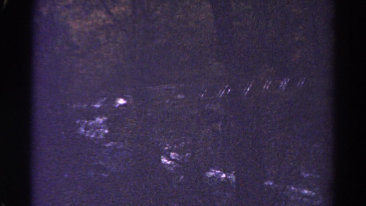 Dark grainy frame with faint river highlights and heavy scratches.