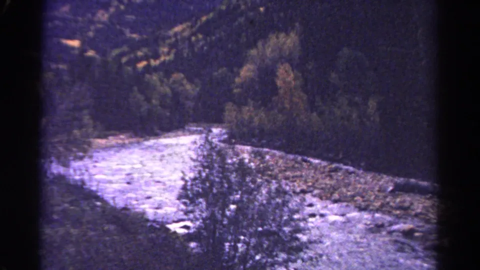 1967 Colorado 8mm view of a pale rushing river bending through a forested mountain valley with rocky banks and dark tree-covered hills.