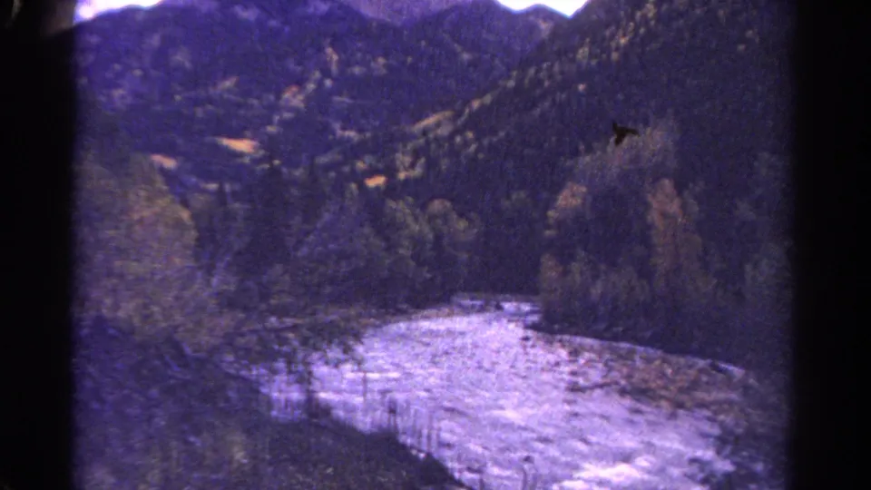 1967 Colorado 8mm frame showing a bright river bend below forested mountain slopes and distant ridges.