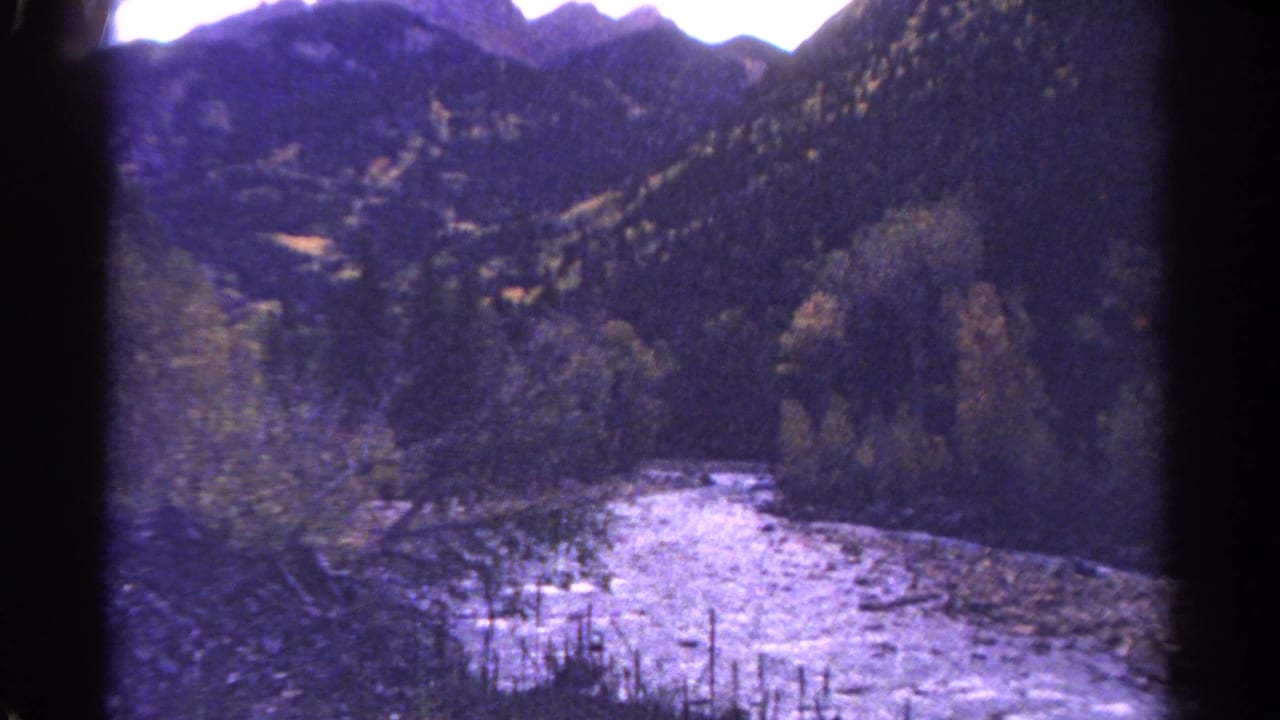 1967 Colorado home-movie frame with a pale river channel running past trees and dark mountain slopes.