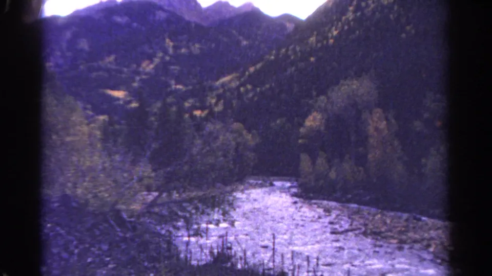 1967 Colorado home-movie frame with a pale river channel running past trees and dark mountain slopes.