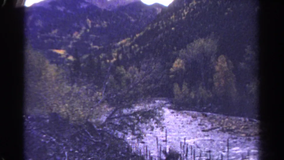 1967 Colorado 8mm frame showing a rocky river edge, foreground trees, and wooded mountain terrain.