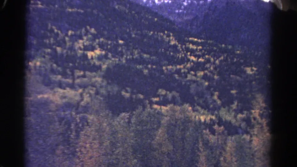 1967 Colorado home-movie frame with a wide wooded valley, ridgelines, and autumn color seen during a moving vehicle shot.