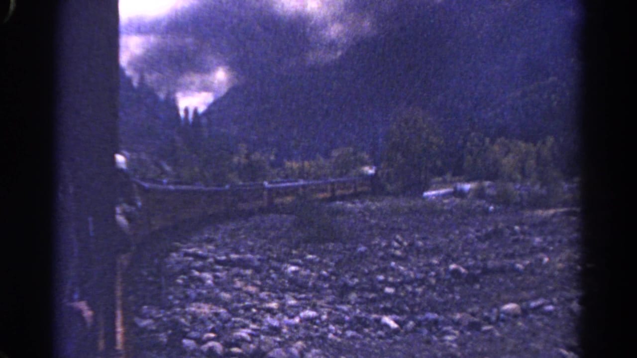 1967 Colorado 8mm frame showing a rocky riverbed, rail line, evergreen slope, and clouded mountain beyond a dark window edge.