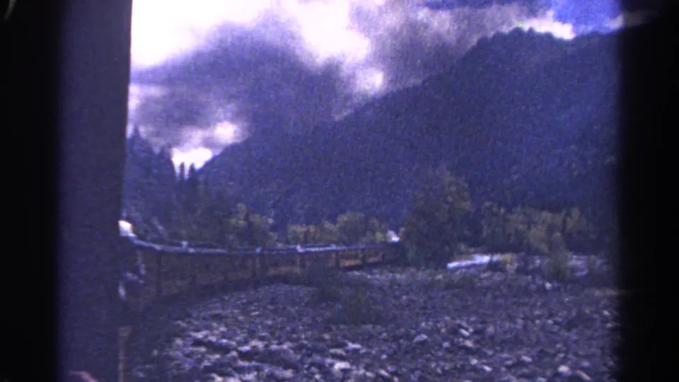 1967 home-movie frame from Colorado showing cloud-covered peaks, forested slopes, a rail line, and a rocky riverbed from a train perspective.
