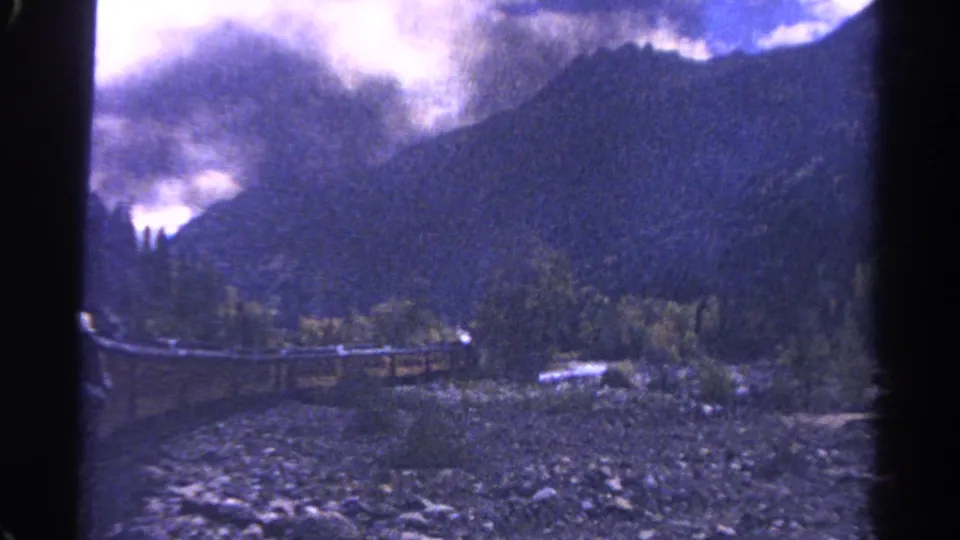 Colorado 8mm travel frame with a train-window edge, rocky foreground, evergreen trees, a rail line, and misty mountain backdrop.