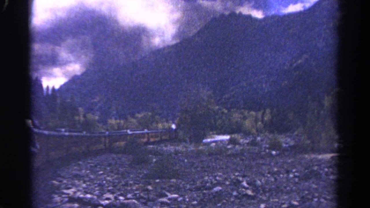 1967 Colorado 8mm frame showing a mountain canyon, cloudy sky, trees, rocky foreground, and a rail or bridge structure near the creek.
