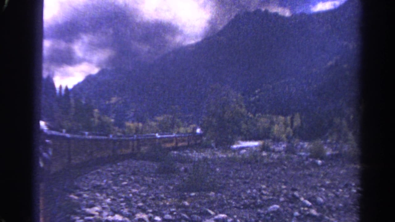 1967 Colorado home-movie view of a mountain valley with wooded slopes, a rocky creek bed, and rail infrastructure through the canyon.