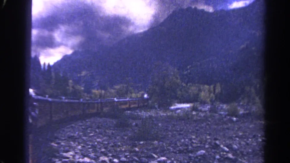 1967 Colorado home-movie view of a mountain valley with wooded slopes, a rocky creek bed, and rail infrastructure through the canyon.