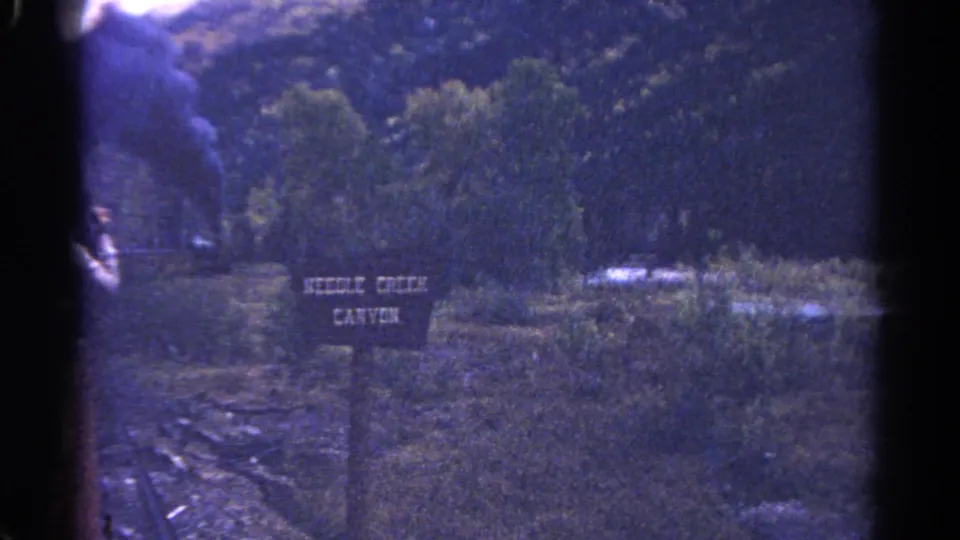 1967 Colorado 8mm frame with a wooden Needle Creek Canyon sign near a rocky rail-side area and wooded mountain backdrop.