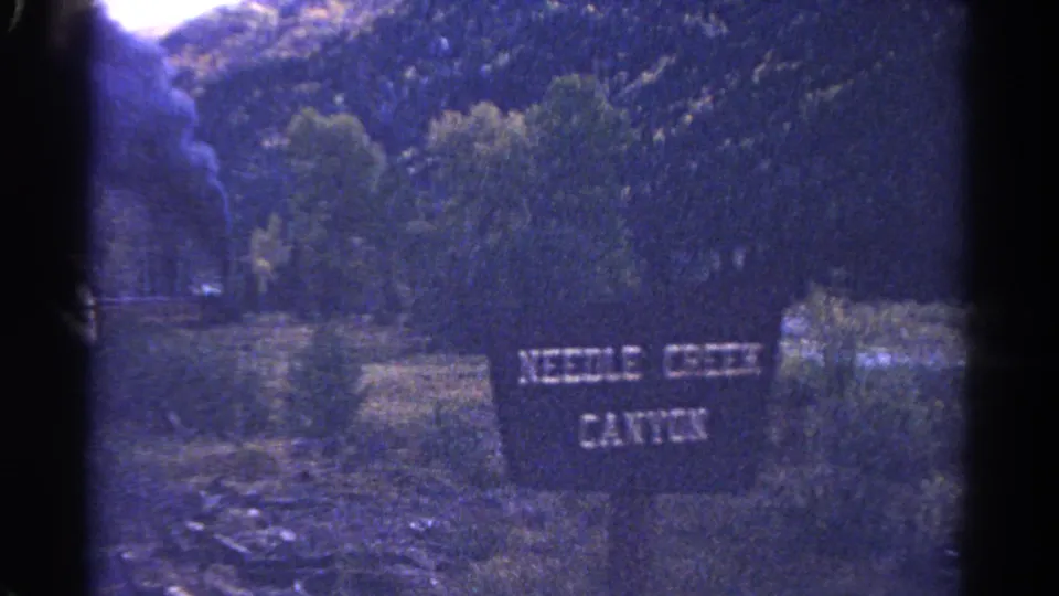 Trackside Needle Creek Canyon sign in a 1967 Colorado 8mm frame, with brush, trees, rocky terrain, and canyon mountains behind it.