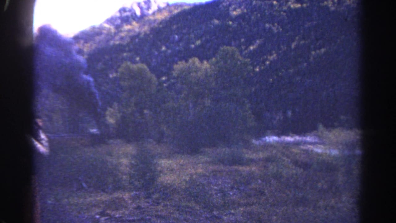 1967 Colorado 8mm train-view frame showing wooded mountains, bright sky, and a plume of dark steam-train smoke.
