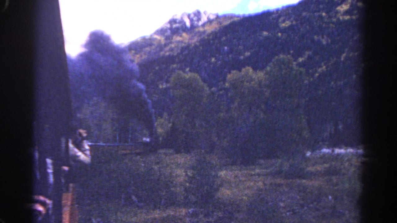 Passenger figure near the side of a train with smoke and mountains beyond.