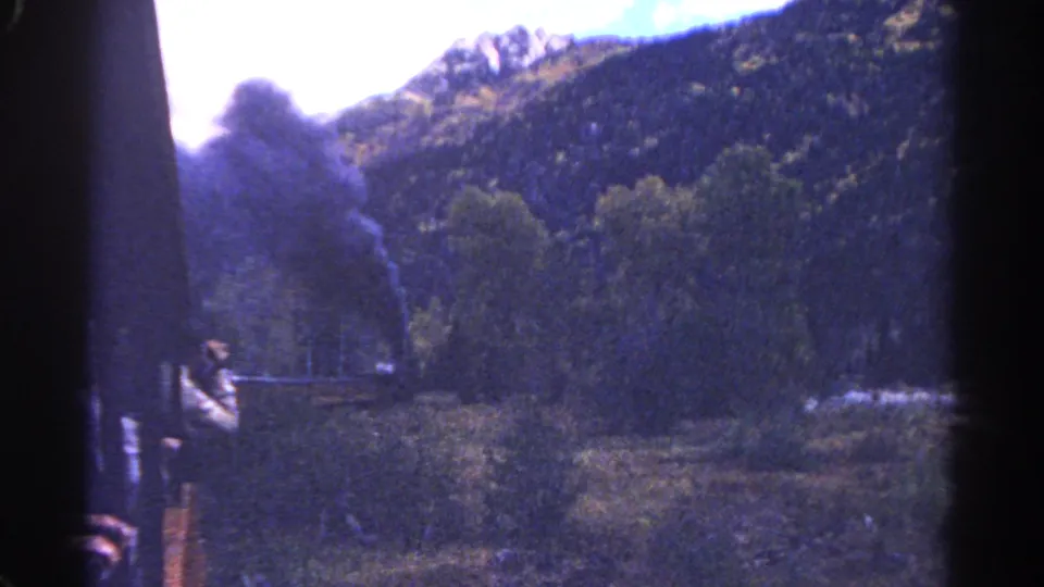 1967 Colorado home-movie frame with a passenger near the train side, dark smoke, trees, and mountain ridges in view.
