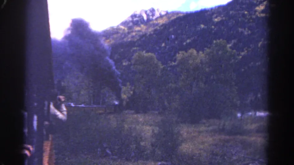 8mm Colorado rail-travel frame showing the train edge, smoke, brush, trees, and a sunlit mountain ridge.