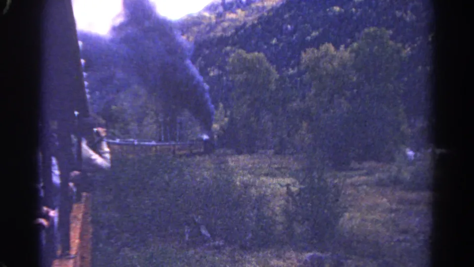 1967 Colorado train-view frame with a passenger near the rail, dark smoke, green trees, and rocky mountain slopes.