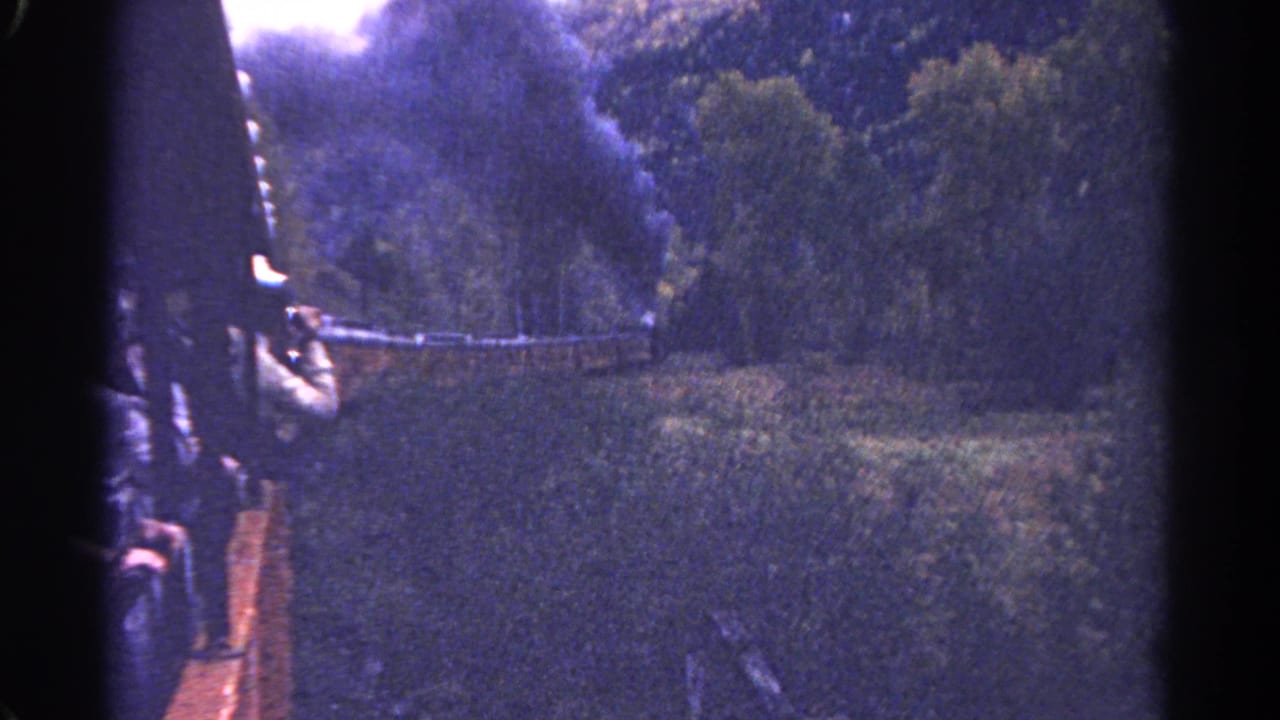 1967 Colorado home-movie frame with an open-air train car, dark smoke, and wooded mountain scenery.