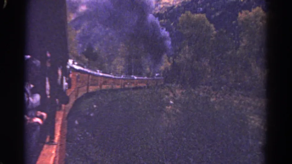 1967 Colorado 8mm still with the train bending through a wooded area, dark smoke above the locomotive and people partly visible at the side.