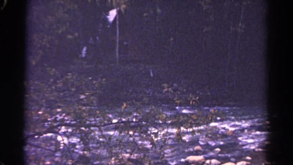 1967 Colorado 8mm frame of a dark forest creek, with rocks in the foreground and a faint pale marker among the trees.