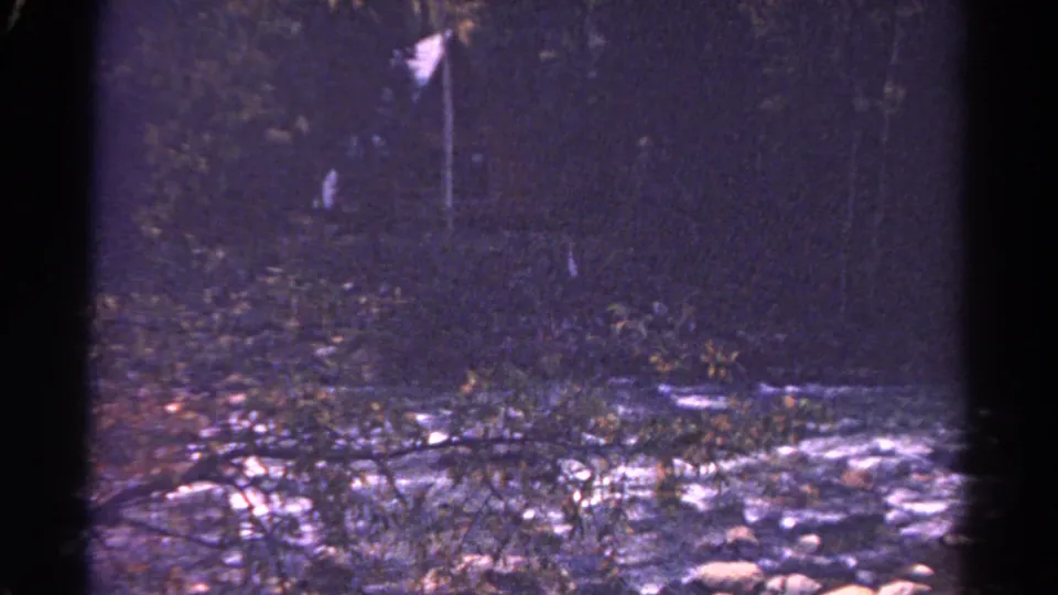 1967 Colorado 8mm frame showing a softly focused dark creek scene with branches, stones, and a distant pale marker.