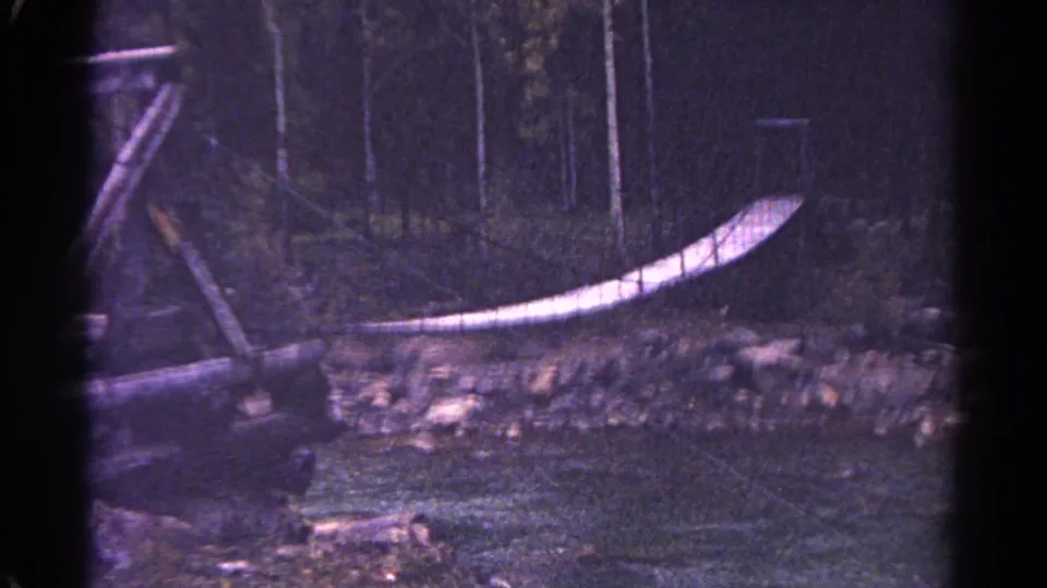 1967 Colorado 8mm frame showing a narrow rope-railed bridge over a stream in the woods.