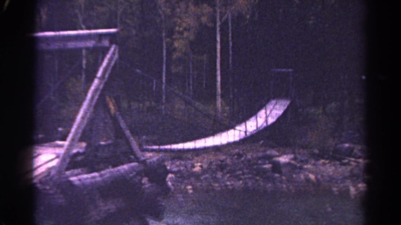 Rustic wooden rope bridge beside rocks and trees in a dim forest setting.