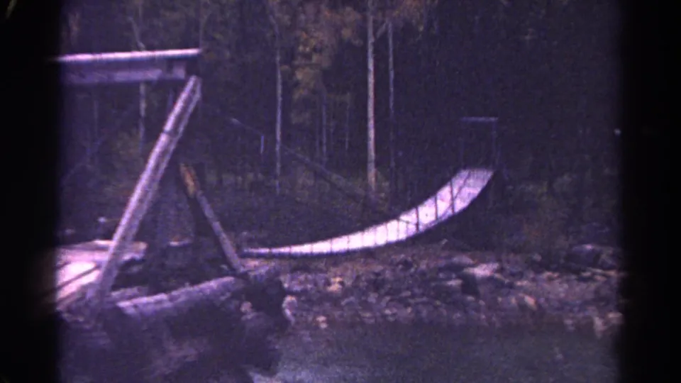 1967 Colorado 8mm frame showing a handmade wooden bridge with angled posts, rope railing and rocky forest ground.