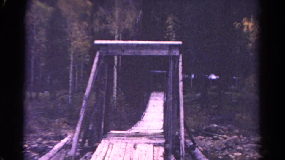 1967 Colorado home-movie frame looking straight along the plank bridge through its wooden support frame in a wooded setting.