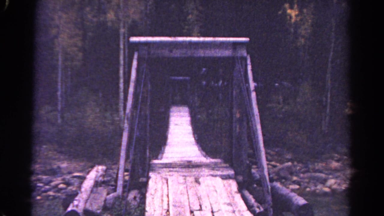 1967 Colorado 8mm frame showing a rustic plank bridge with rope railings leading into a mountain forest.
