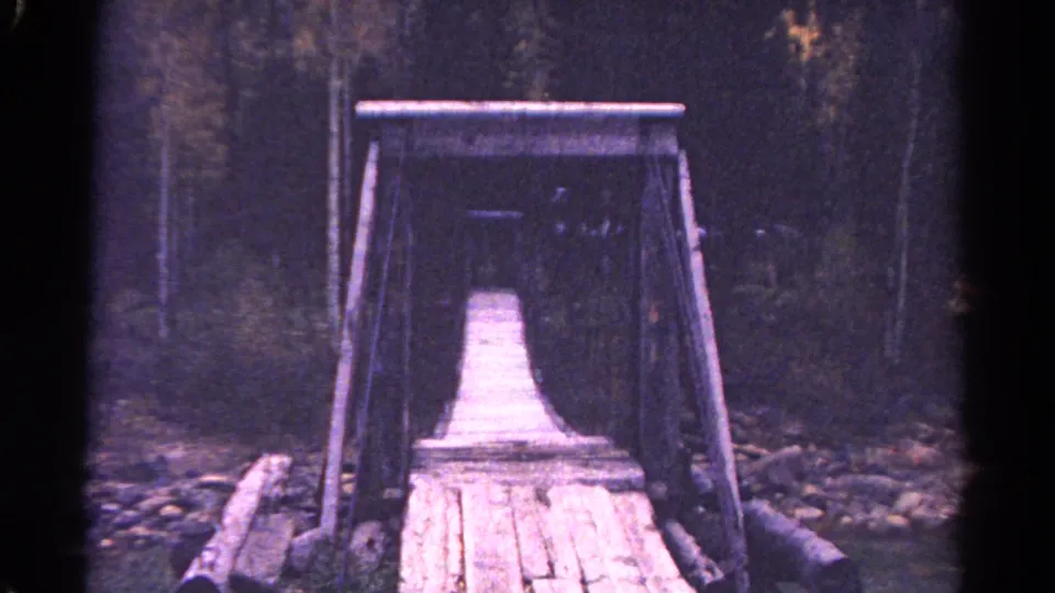 1967 Colorado 8mm frame showing a rustic plank bridge with rope railings leading into a mountain forest.