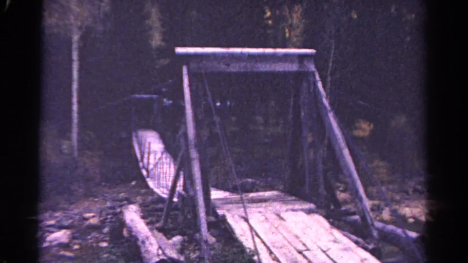 A 1967 Colorado mountain bridge is seen from an angle, with rope railings, log framing, and dense woods beyond.