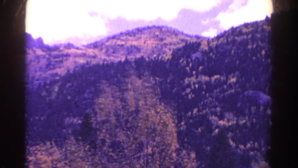A 1967 Colorado home-movie frame looks across a mountain valley with evergreen-covered slopes and a light path through the terrain.