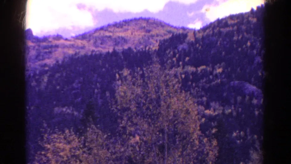 1967 Colorado 8mm frame showing a wooded mountain ridge under a bright cloudy sky with purple-blue film fading.