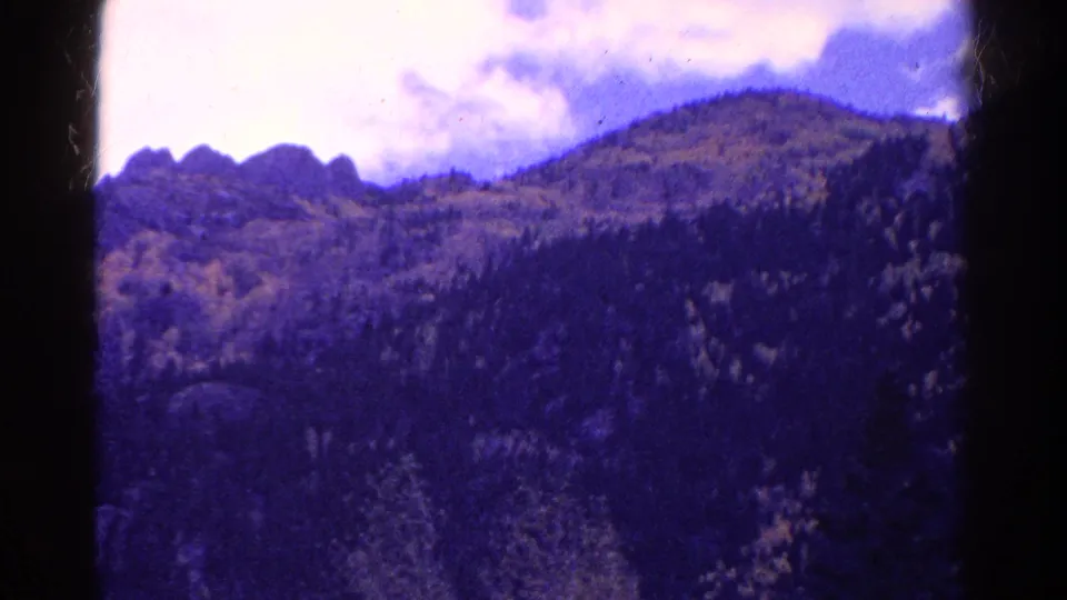 1967 Colorado 8mm frame of a forested mountain landscape with distant rocky peaks, bright clouds, and aged color shift.