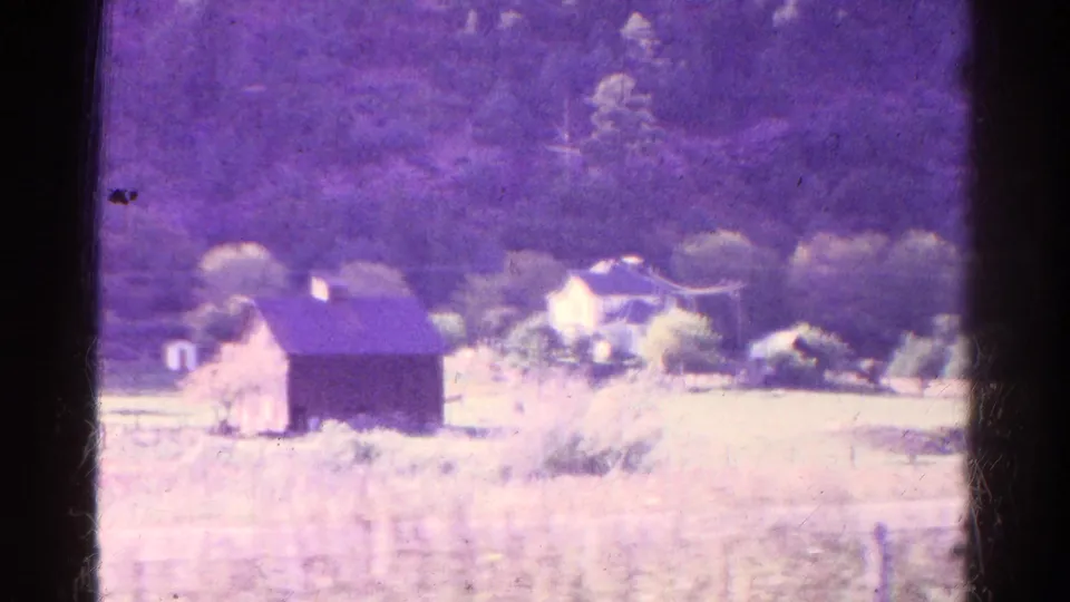 1967 Colorado countryside frame showing an open field, barn-like building, distant house, and wooded mountain backdrop.