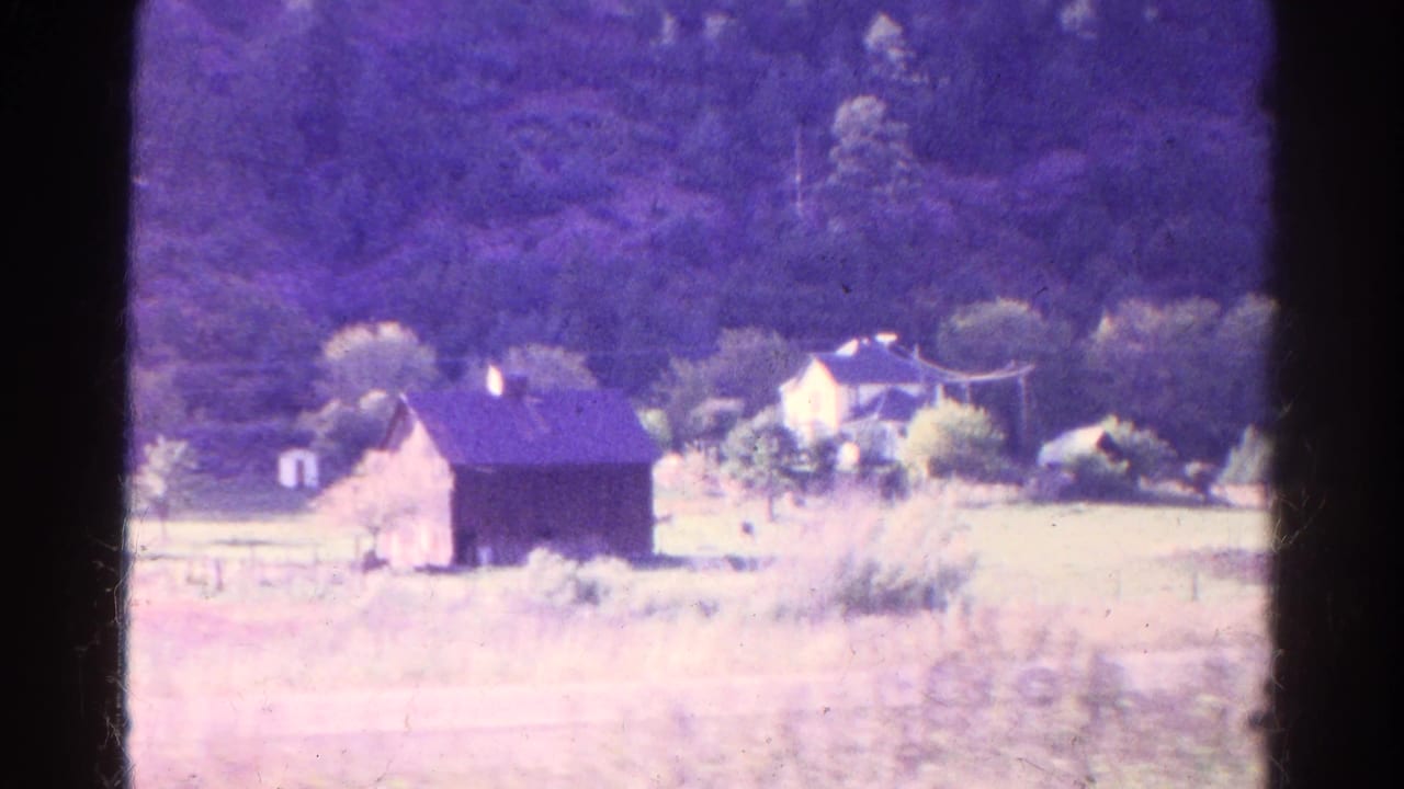 Distant house and barn in a grassy field below a wooded hillside.
