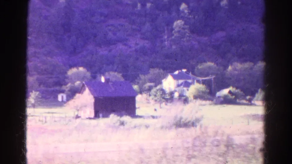 1967 Colorado home-movie frame showing a rural house, barn, open field, fence line, and wooded hillside with aged color shift.