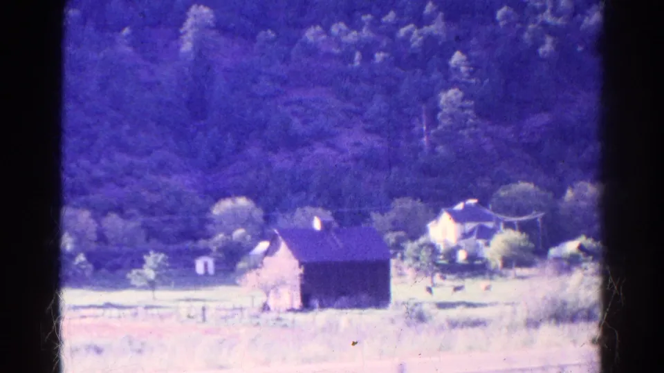 Aged 8mm frame of a rural Colorado house and barn area, with fence posts, field grass, bushes, and a dark wooded hillside.