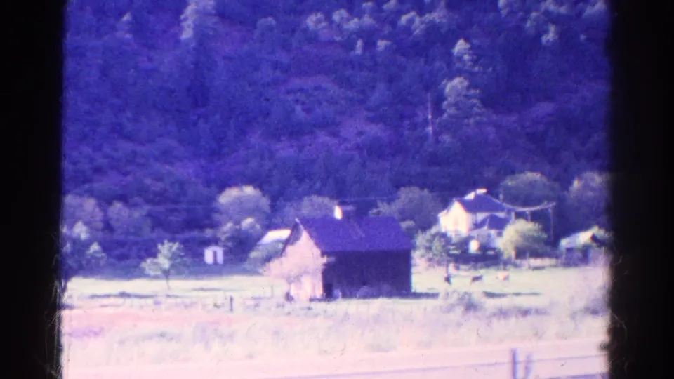 1967 Colorado 8mm still showing a rural house, barn, pale open field, fence line, and forested hill with magenta-blue film shift.