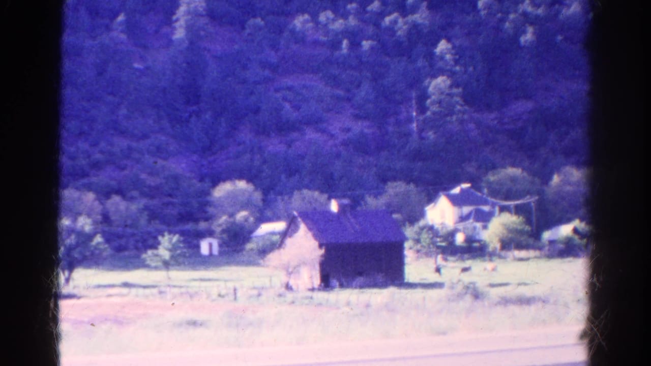 Rural farmstead with a dark barn, house, trees, pasture, and distant wooded hillside seen through a train window.