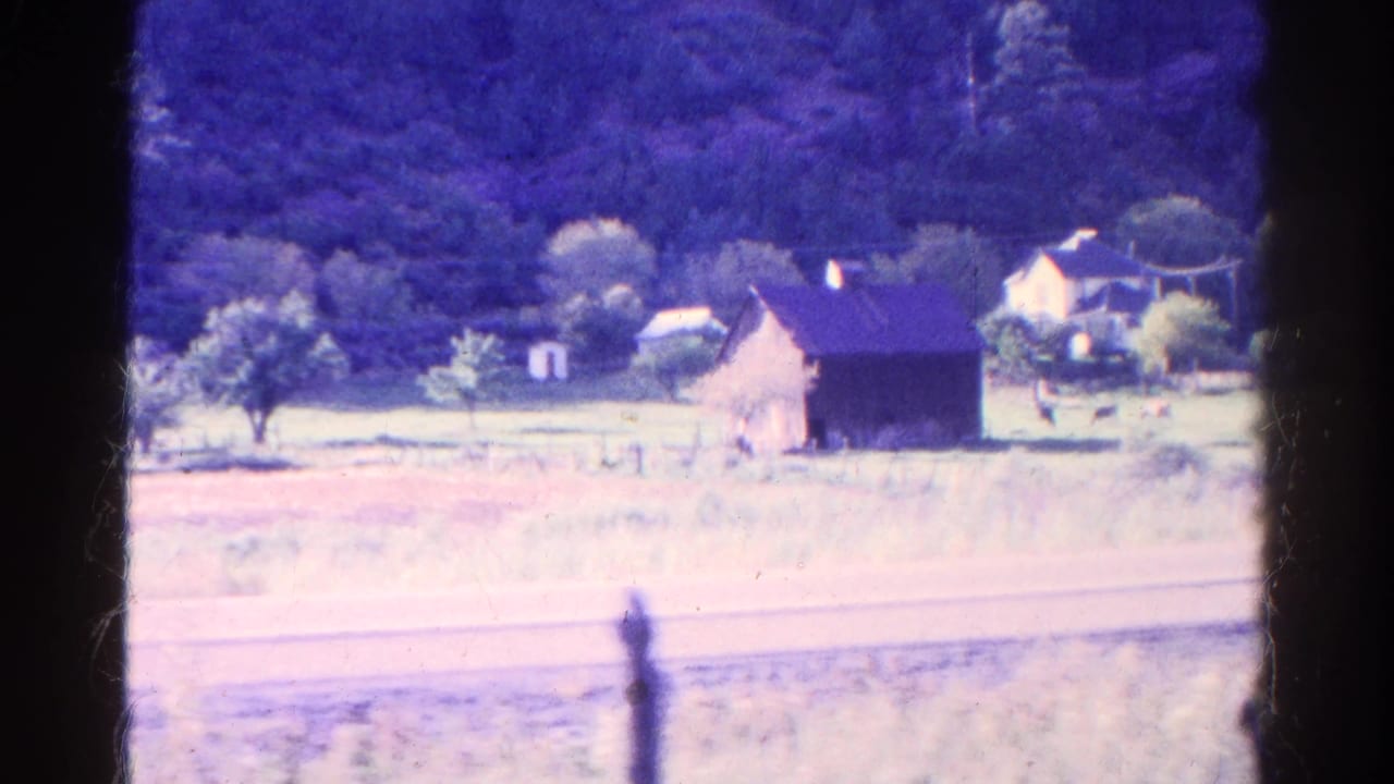 Farm buildings and trees beside open pasture, framed by dark train-window edges and a distant hillside.