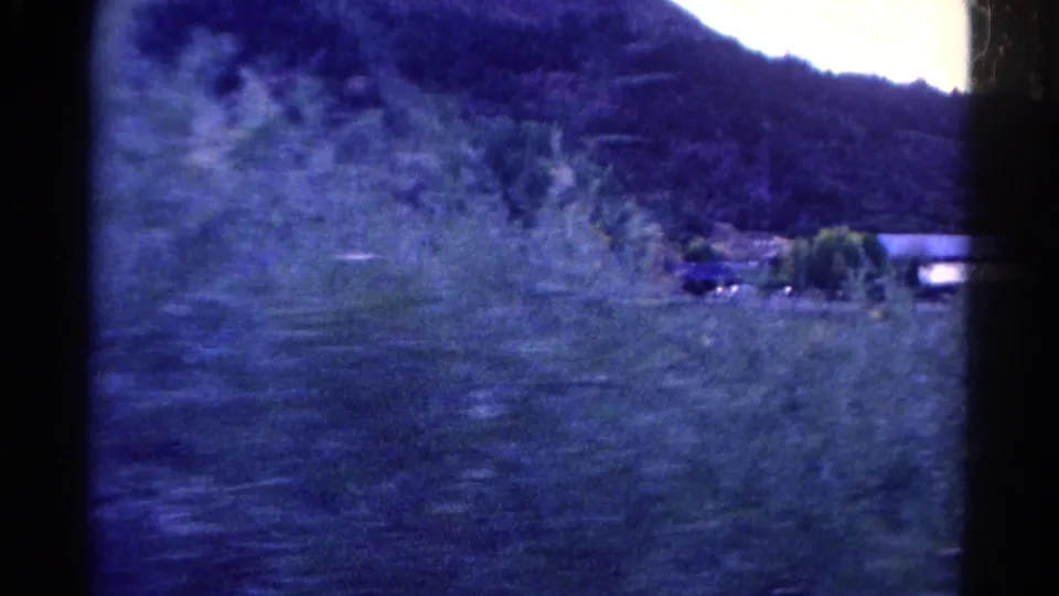 1967 Colorado 8mm frame with a light-clad person partly visible in tall grass, facing a rural hillside and distant buildings.