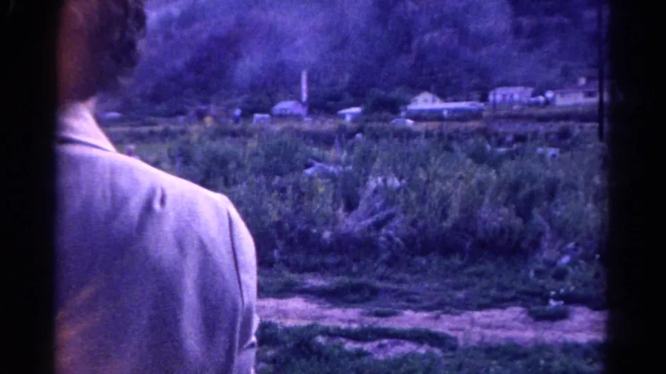 1967 Colorado 8mm frame showing a light-jacketed passenger at a vehicle window with rural brush and small houses beyond.