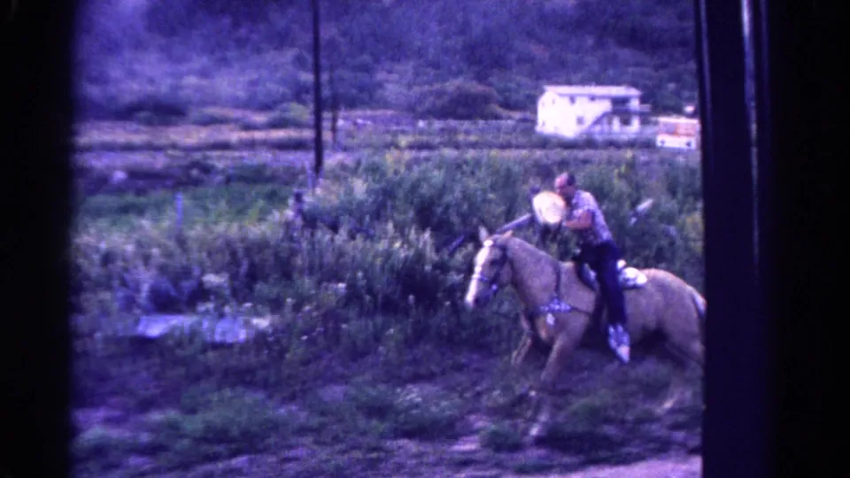 1967 Colorado home-movie frame showing a palomino horse and rider in a field below utility poles and a distant white building.